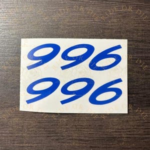 May include: A rectangular white sticker sheet featuring two rows of the number 996 printed in a bold, blue, sans-serif font. The numbers are arranged with one set of 996 above the other on a plain, light-colored background surface.