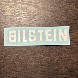 May include: A rectangular sticker with the word "BILSTEIN" in white capital letters. The sticker has a light blue background and is placed on a brown wooden surface.