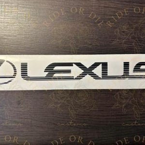 May include: A white Lexus decal with black lettering and logo. The Lexus logo is a stylized letter "L" inside an oval. The word "LEXUS" is written in a bold, sans-serif font.