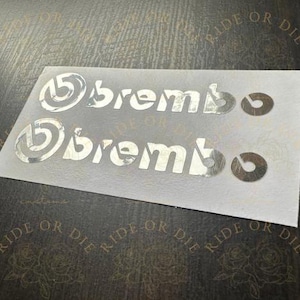 May include: Two silver metallic adhesive decals featuring the Brembo logo and brand name arranged in two horizontal rows on a white backing sheet. Each row includes the circular logo icon followed by the text brembo and a small circular dot.