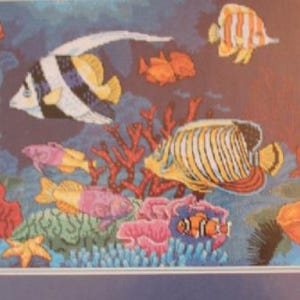 May include: A colorful cross-stitch depicting an underwater scene. The artwork features various fish, including a black and white striped fish, and coral in shades of red, pink, and orange. The background is a gradient of blues and browns.