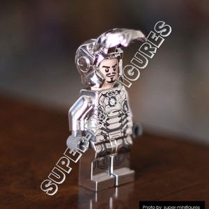 Puede incluir: Una minifigura Lego de Iron Man plateada con un traje plateado y negro y un casco plateado. La minifigura est&aacute; de pie sobre una superficie de madera.