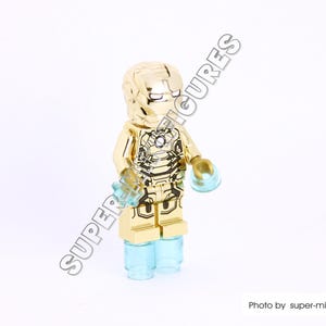 Puede incluir: Minifigura Lego de Iron Man de color dorado con una base azul transl&uacute;cida. La minifigura tiene un traje dorado detallado con detalles negros y una cara roja.