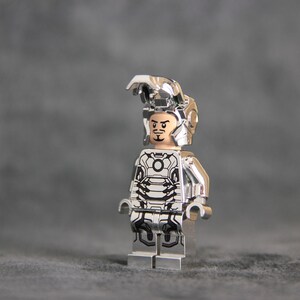 Puede incluir: Una minifigura Lego plateada de Iron Man con un traje plateado con un dise&ntilde;o en blanco y negro. La minifigura tiene una cara negra con una expresi&oacute;n seria.
