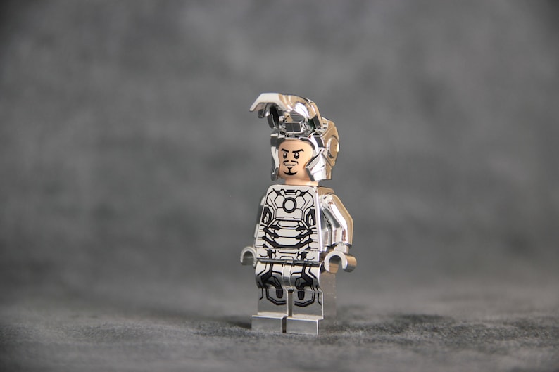 Puede incluir: Una minifigura Lego plateada de Iron Man con un traje plateado con un dise&ntilde;o en blanco y negro. La minifigura tiene una cara negra con una expresi&oacute;n seria.