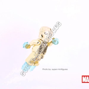 Puede incluir: Minifigura Lego dorada de Iron Man con una pieza de pecho transl&uacute;cida azul claro y una mano transl&uacute;cida azul claro. La minifigura est&aacute; en pose con un brazo levantado y el otro brazo extendido. La minifigura est&aacute; sobre un fondo blanco. Foto de super-minifigures. MARVEL