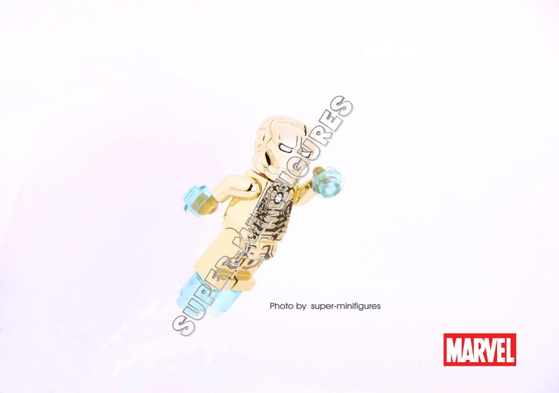 Puede incluir: Minifigura Lego dorada de Iron Man con una pieza de pecho transl&uacute;cida azul claro y una mano transl&uacute;cida azul claro. La minifigura est&aacute; en pose con un brazo levantado y el otro brazo extendido. La minifigura est&aacute; sobre un fondo blanco. Foto de super-minifigures. MARVEL
