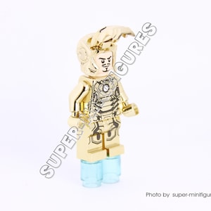 Puede incluir: Minifigura Lego de Iron Man de color dorado con un casco y una armadura dorados con detalles negros. La minifigura est&aacute; de pie sobre una base azul claro.