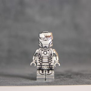 Puede incluir: Una minifigura Lego de Iron Man plateada con un dise&ntilde;o impreso en blanco y negro en el pecho y la cabeza. La minifigura tiene un casco plateado con una visera negra y un cuerpo plateado con detalles negros.
