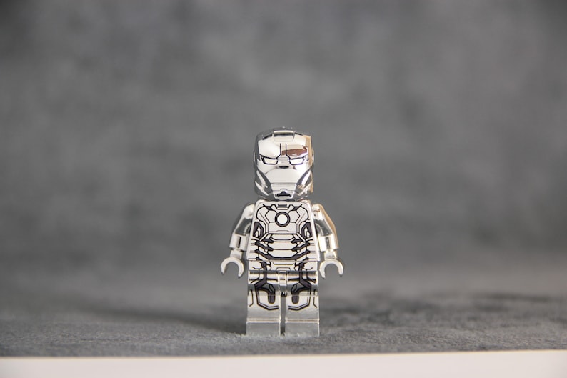 Puede incluir: Una minifigura Lego de Iron Man plateada con un dise&ntilde;o impreso en blanco y negro en el pecho y la cabeza. La minifigura tiene un casco plateado con una visera negra y un cuerpo plateado con detalles negros.
