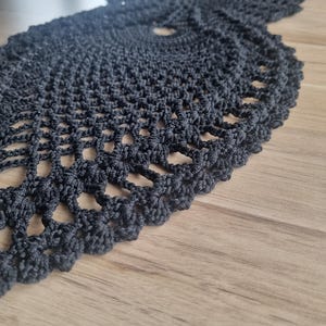 Könnte beinhalten: Handgefertigter, schwarzer Häkeltischläufer mit detailliertem Lochmuster. Der Läufer hat einen gewellten Rand und ein kreisförmiges Muster in der Mitte. Das filigrane Design ist auf der Holzoberfläche sichtbar.