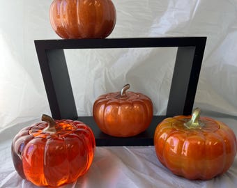May include: Four orange decorative pumpkins with stems. One pumpkin is on a black shelf, and the other three are on a white surface. The pumpkins have a glossy finish and are in various shades of orange.