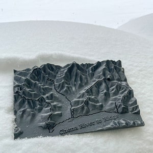 May include: A 3D printed model of the Chena River to Ridge trail in Alaska. The model is made of gray plastic and shows the terrain of the trail. The model is sitting on a bed of snow.