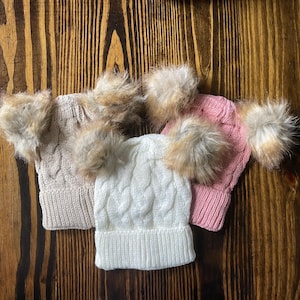 Könnte beinhalten: Drei Zopfstrickmützen mit Kunstfellbommeln. Eine ist beige, eine ist weiß und eine ist rosa. Die Mützen sind auf einer dunklen Holzoberfläche angeordnet, die ihre Textur und ihr Design zeigt.