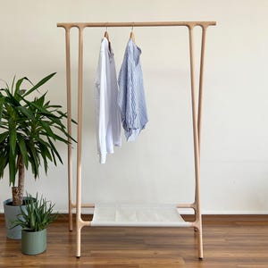 May include: A light-coloured wooden clothing rack with four vertical legs and a horizontal top bar. A white long-sleeve shirt and a blue and white striped long-sleeve shirt hang from wooden hangers. A white fabric shelf is positioned at the base.