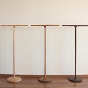 Puede incluir: Tres soportes de madera en forma de T, cada uno con una base circular, un poste vertical y una barra horizontal superior. Los soportes se presentan en tres tonos de madera distintos: un acabado natural claro, un marrón medio y un marrón oscuro.