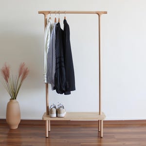 Peut inclure: Un portant à vêtements en bois clair doté d'une barre de suspension horizontale et d'une étagère inférieure. Quatre cintres portent une chemise rayée blanche, une chemise rayée bleue et deux vêtements noirs. Une paire de baskets blanches est posée.