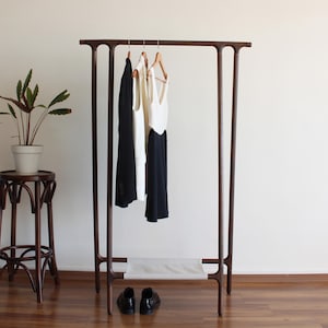 Peut inclure: Un portant à vêtements en bois brun foncé avec une barre de suspension horizontale et une étagère inférieure en tissu clair. Trois cintres en bois portent une veste noire, un haut sans manches blanc et une robe noire et blanche. Des chaussures noires.