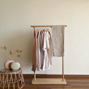 May include: A light-coloured wooden clothing rack holds several shirts in shades of white, beige, and dusty rose, along with a folded beige fabric piece. Beside the rack, two smooth, rounded ceramic vases sit on a small, round rattan table.