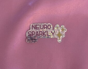 Puede incluir: Una pegatina transparente y brillante con las palabras "NEURO SPARKLY" en letras moradas. La pegatina tiene una forma única con dos estrellas amarillas en el lado derecho. El fondo es una tela de satén rosa.