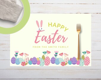 May include: A cream-colored Easter placemat with the words "HAPPY Easter" in pink and green, and "FROM THE SMITH FAMILY" below. A bunny graphic and floral and egg border complete the design. A gold fork is on the right.