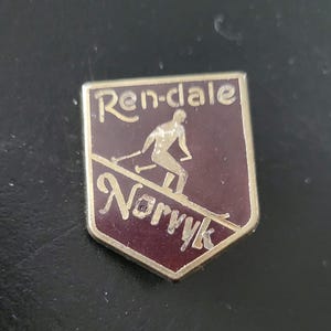 May include: A vintage metal pin with a maroon background and gold lettering. The pin features a skier on a slope with the words "Ren-dale" and "Norrvik" in gold lettering.