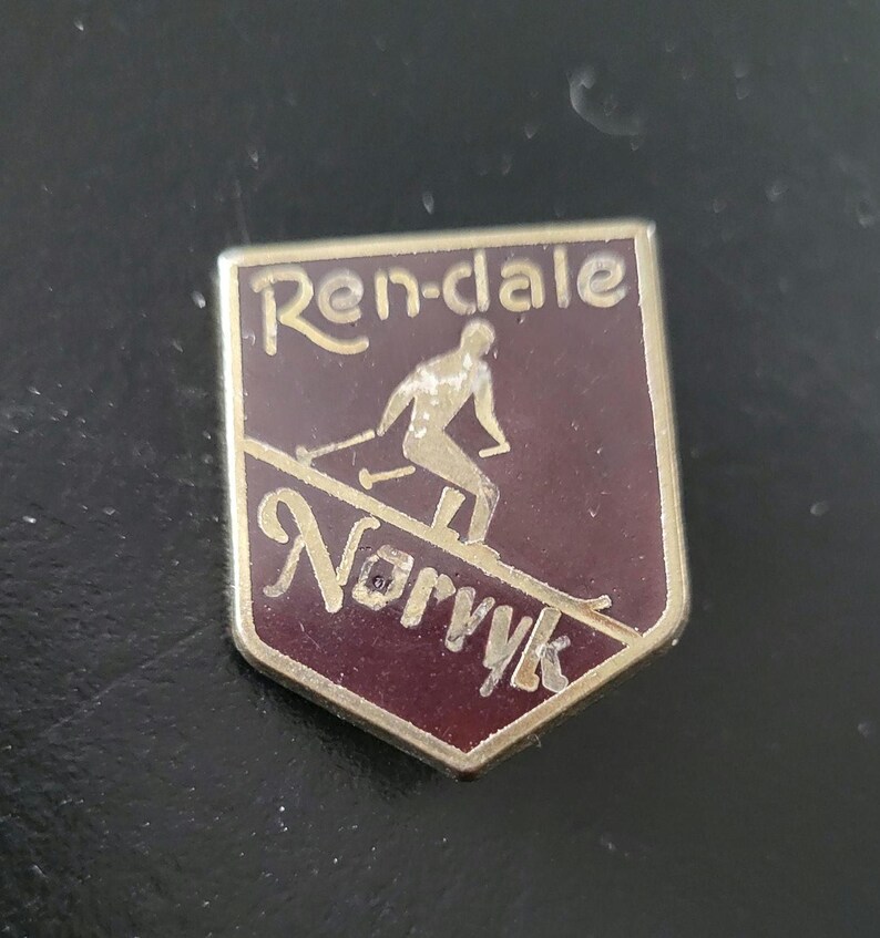 May include: A vintage metal pin with a maroon background and gold lettering. The pin features a skier on a slope with the words "Ren-dale" and "Norrvik" in gold lettering.