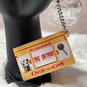 May include: A yellow keychain shaped like a vintage VHS tape featuring the text HALLOWEEN NIGHT, TRIC OR TREAT, and CHECK YOUR CANDY. The design includes a small illustration of a cat and a lollipop with a jack-o'-lantern face on a metal chain.