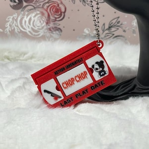 May include: A red rectangular keychain shaped like a VHS tape with a silver ball chain. The label features the text "RETURN IMMEDIATELY," "CHOP CHOP," and "LAST PLAY DATE," along with a graphic of a knife and a black and white bear character.