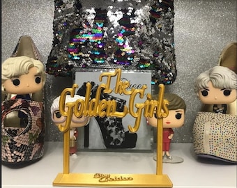 May include: Gold-colored decorative stand with the words "The Golden Girls" in a script font. The stand features a small picture of a high-heeled shoe. There are three small figurines of the Golden Girls characters on the stand.