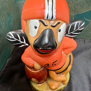 May include: A ceramic figurine of a cartoon football player wearing an orange helmet with white stripes and a red jersey with the number 85. The figurine is holding a brown football and has wings with black and white markings. The base of the figurine is green and says "19 GNAT"
