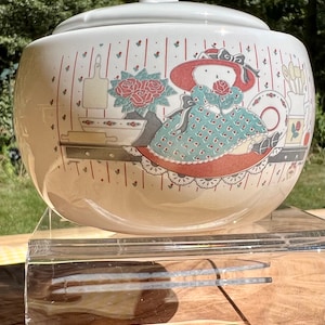 May include: A white ceramic crock with a lid, featuring a whimsical illustration of a girl in a red hat and a green dress, holding a red rose. The crock has a red and white striped pattern and a floral design.