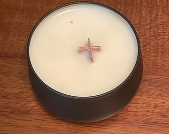 Puede incluir: Un tarro de vela negro con una vela de cera blanca y una cruz de madera en la parte superior. La vela está en un tarro negro con un acabado mate.