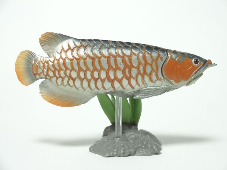 May include: A detailed fish figurine in silver and orange. The fish has orange fins and head, with a silver body featuring an orange scale pattern. It is mounted on a clear stand with green plastic plants and a grey base.
