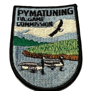May include: An embroidered patch with a blue background and a black border. The patch features a scene of a lake with green trees and a flying eagle. The text "PYMATUNING PA GAME COMMISSION" is embroidered in black on the patch.