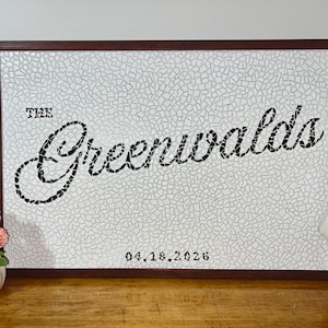 May include: A rectangular wall sign with a dark brown frame featuring a white mosaic tile background. The text "THE Greenwalds" is written in black script, and the date "04.18.2026" is printed in black at the bottom center of the sign.