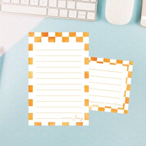 May include: Two white notepads with orange checkered borders and lined pages. The larger notepad has the text "A note from Jenny." Pencils and a keyboard are in the background.