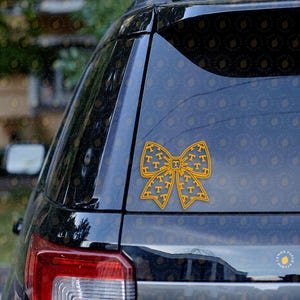 May include: A yellow bow-shaped decal with a repeating "T" logo pattern, affixed to the rear window of a black vehicle. The decal is a decorative accessory, likely for a car or truck, and is designed to be applied to a glass surface.