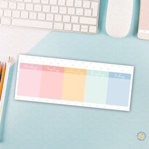 May include: A rectangular weekly planner with pastel-colored sections for each weekday: Monday, Tuesday, Wednesday, Thursday, and Friday. The planner has a white border with small heart patterns. The background is a light blue surface.