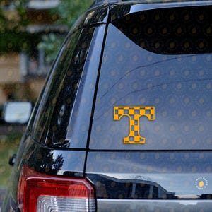 May include: A yellow and black checkered "T" logo sticker is affixed to the rear window of a black vehicle. The sticker is a stylized representation of the letter "T", with a checkered pattern in the center.