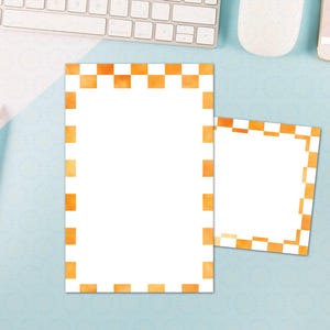 May include: Two white notepads with orange checkered borders. One notepad is rectangular, and the other is square. Pencils and a keyboard are in the background. The notepads are on a light blue surface.
