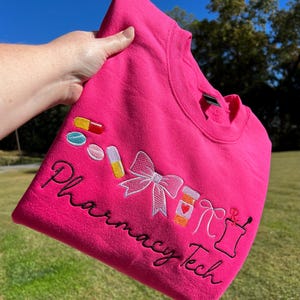 May include: A bright pink sweatshirt with embroidered pharmacy-themed designs. The design includes pills, a bow, a prescription bottle with a heart, and the words "Pharmacy Tech" in cursive.