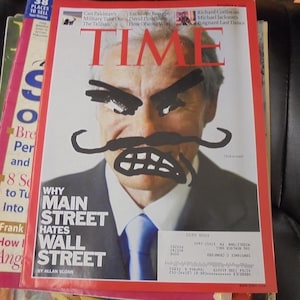 May include: A stack of magazines, with the top one featuring a TIME magazine cover. The cover has a portrait of a man with drawn-on angry eyebrows, a mustache, and teeth. The headline reads "WHY MAIN STREET HATES WALL STREET."