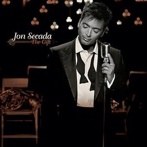 May include: Album cover featuring Jon Secada in a black tuxedo, holding a vintage microphone. The title "The Gift" is displayed with elegant script. The background is a dimly lit stage with musical instruments.