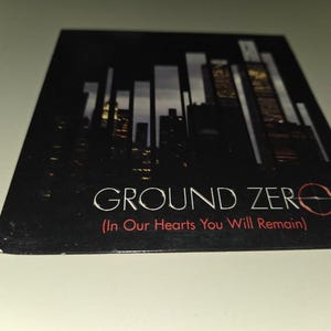 May include: A black CD case featuring a nighttime cityscape silhouette. The words "GROUND ZERO" are in white, with "(In Our Hearts You Will Remain)" in red below. The design evokes a sense of remembrance.