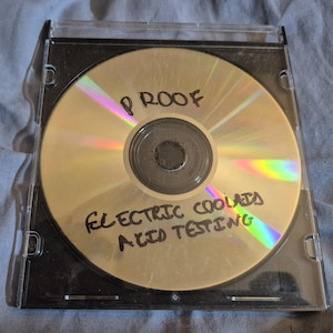 Puede incluir: Un disco compacto en una funda de plástico transparente. El disco dorado tiene texto escrito a mano: "PROOF" y "ELECTRIC COOLAIDS ACID TESTING". La funda está ligeramente polvorienta.