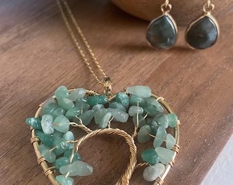 Puede incluir: Un collar de oro con un colgante en forma de corazón que presenta un diseño de árbol de la vida envuelto en alambre con piedras preciosas verdes. Aretes colgantes a juego con piedras preciosas verdes.