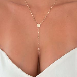 Peut inclure: Collier dor&eacute; avec une cha&icirc;ne d&eacute;licate orn&eacute;e de petites perles. Le collier comporte un pendentif avec une petite croix et un m&eacute;daillon circulaire. Le collier est port&eacute; sur un haut blanc.