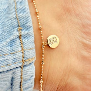 Op de afbeelding: Goudkleurige enkelband met een fijne ketting van kleine kralen en een bungelende bedel. De bedel is een ronde schijf met de letter "M" ingelegd met kleine, glinsterende steentjes. De enkelband wordt op een enkel gedragen.