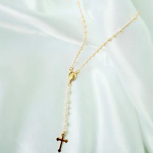 Peut inclure: Un collier de chapelet dor&eacute; avec un pendentif en forme de croix et un petit pendentif ovale repr&eacute;sentant une figure religieuse. Le collier est compos&eacute; de petites perles rondes et est pr&eacute;sent&eacute; sur un tissu blanc et soyeux.